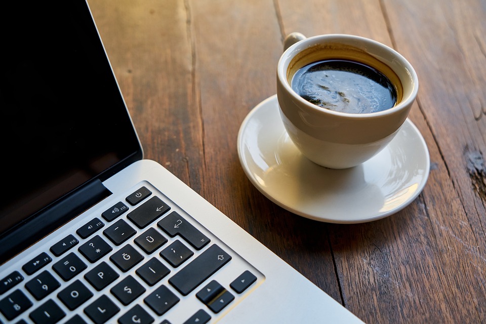 Partial view of a laptop keyboard next to a cup of black coffee on a saucer