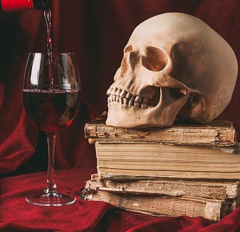 A table with a red velvet cloth holding a glass of red wine, being filled, a pile of old books on top of which is a skull